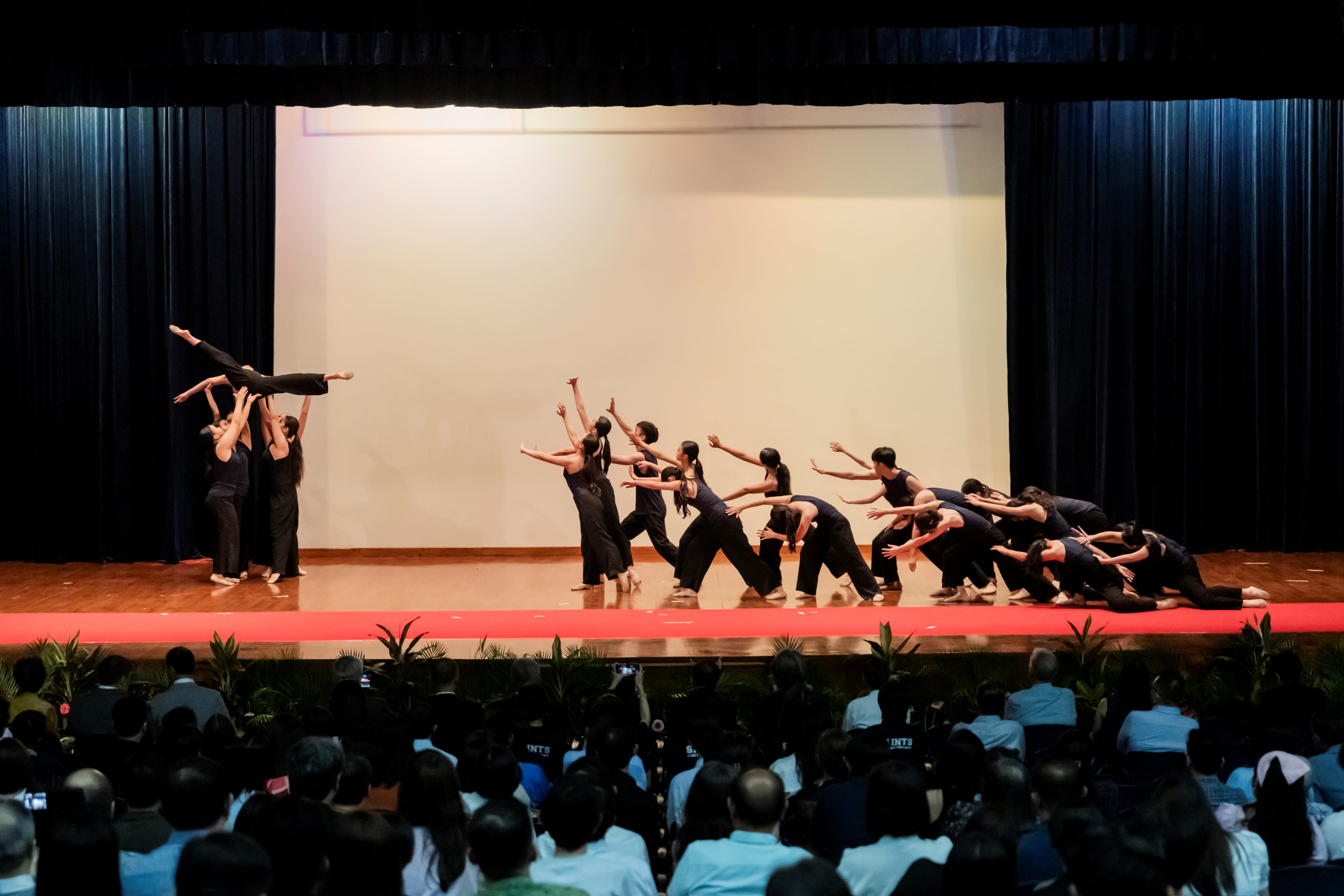 A group of dancers in dark outfits performing a contemporary dance routine on stage, featuring an overhead lift, watched by a seated audience.
